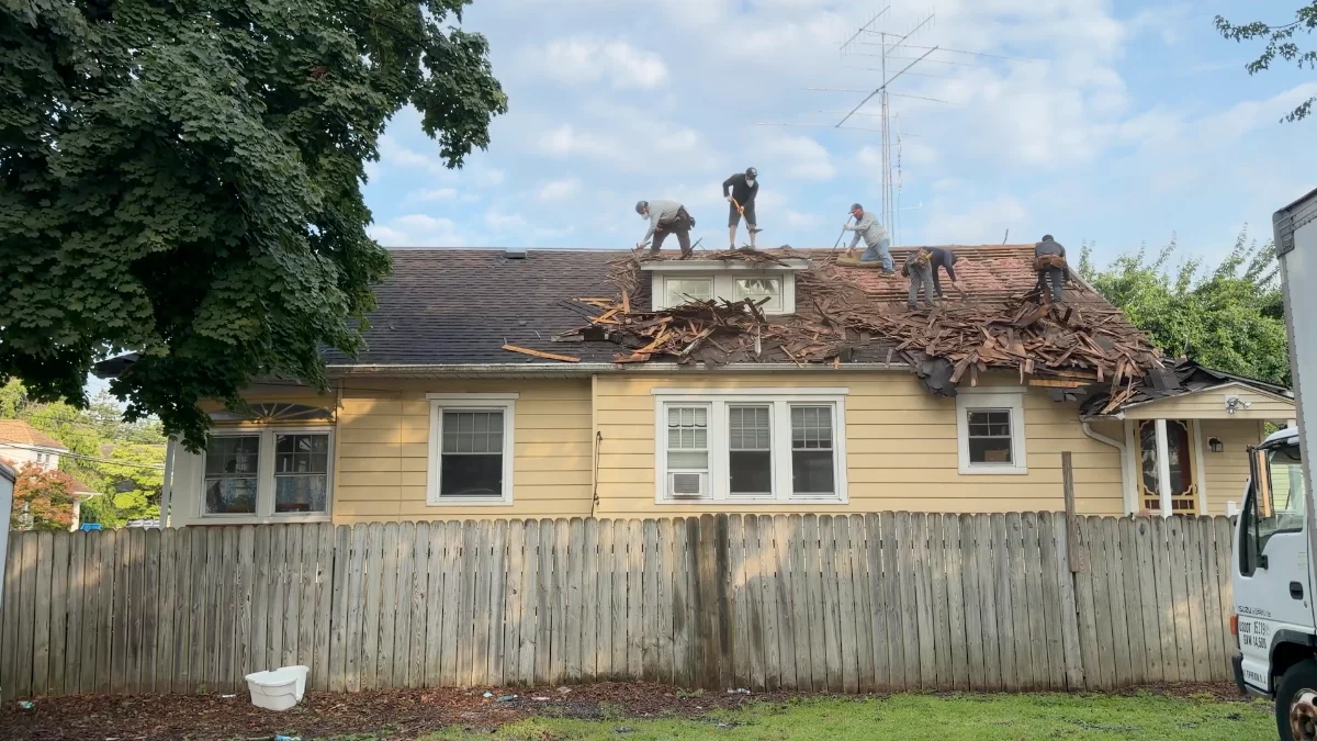 Five roofing crew members removing layered roofing and cedar shake from a one-story home with spaced wood roof boards exposed